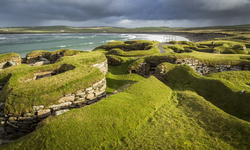 View of the neolithic village Skara Brae. Low stone walls are topped by turf. The sea is in the background.