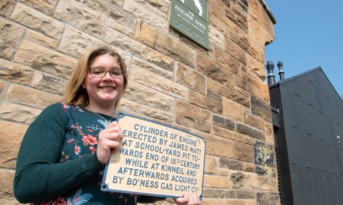 A person stands outside the Engine Shed smiling, holding up a plaque