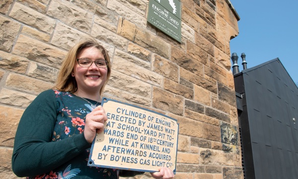 A person stands outside the Engine Shed smiling, holding up a plaque