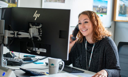 A woman looks at a computer screen, smiling while speaking on the phone.