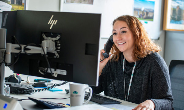 A woman looks at a computer screen, smiling while speaking on the phone.