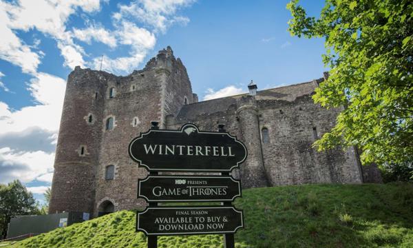 Doune Castle on a sunny day with a sign saying "Winterfell" in front of it.