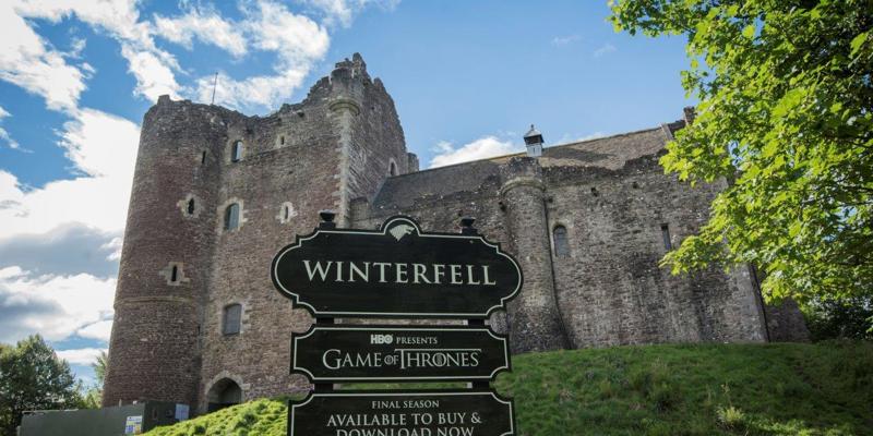 Doune Castle on a sunny day with a sign saying "Winterfell" in front of it.