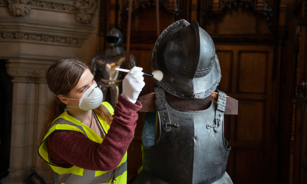 A conservation apprentice works on a set of armour with a fine brush.