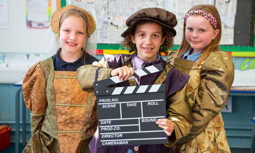 Three pupils from from Bun-sgoil Taobh na Pàirce, Edinburgh pose with a director's clapperboard. They are wearing costumes inspired by the fashion of the 16th-century.