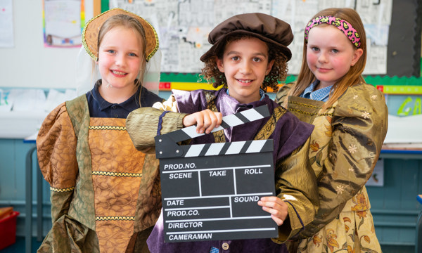 Three pupils from from Bun-sgoil Taobh na Pàirce, Edinburgh pose with a director's clapperboard. They are wearing costumes inspired by the fashion of the 16th-century.