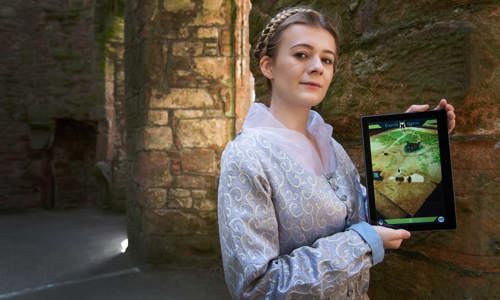 A woman dressed in a lilac, historic dress stands holding an iPad.