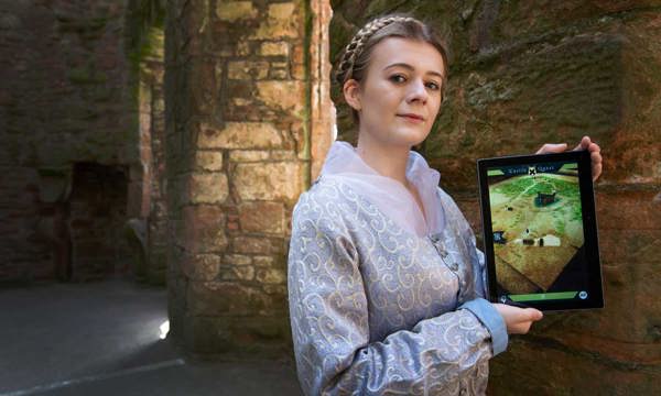 A woman dressed in a lilac, historic dress stands holding an iPad.