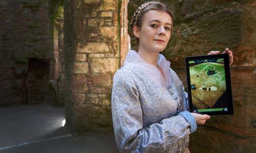 A woman dressed in a lilac, historic dress stands holding an iPad.