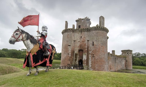 A knight in full armour poses in front of a castle on a white horse dressed in red and black colours 