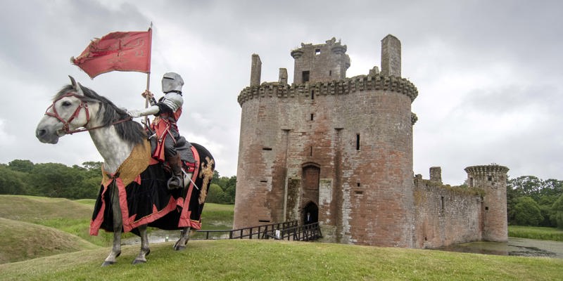 A knight in full armour poses in front of a castle on a white horse dressed in red and black colours 