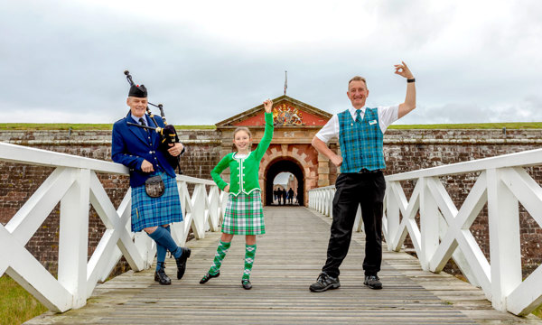 A young girl in a green kilt dances with a steward and piper in front of fort george