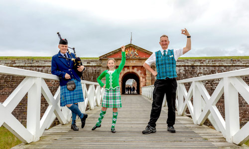 A young girl in a green kilt dances with a steward and piper in front of fort george