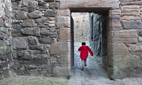 A small boy in a red coat runs excitedly through a doorway in a medieval castle 