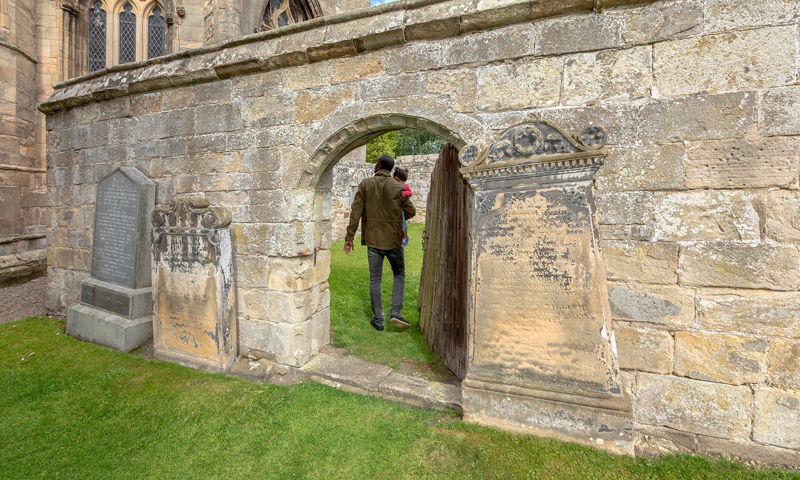 A man carrying a child walks through an ancient doorway while exploring the ruins of an abbey 
