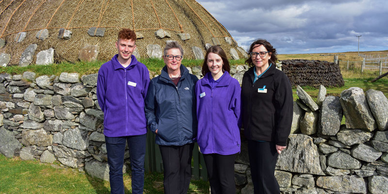 Four people stand in front of a thatched roof building. Two are wearing purple fleeces.