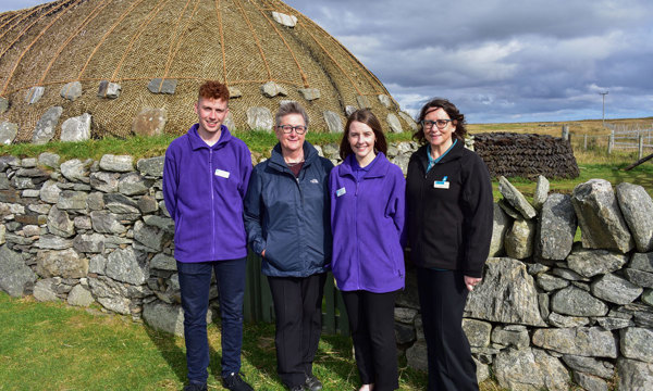 Four people stand in front of a thatched roof building. Two are wearing purple fleeces.