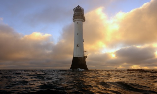 A white lighthouse standing alone in a choppy sea as sun breaks through clouds in the background. Seabirds are perched on the base of the lighthouse. 
