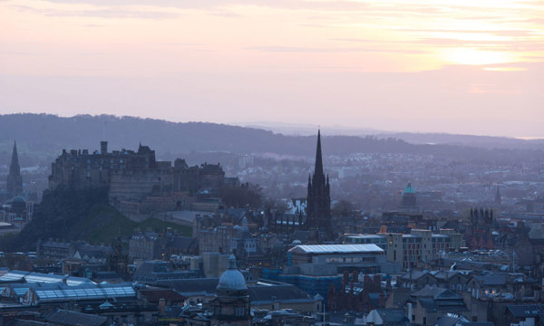 A cityscape at sunset with the outline of a castle on a hill and spire of a church rising above the city.