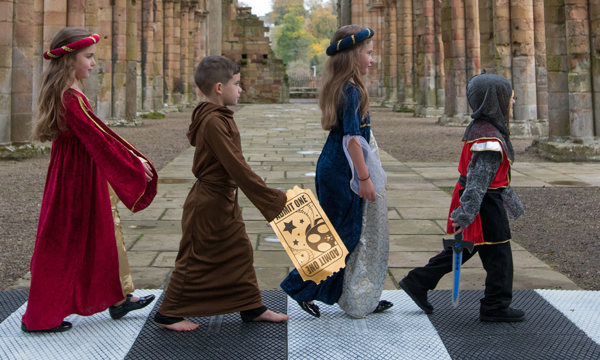 Image of a group of young kids walking across 'Abbey Road' dressed as medieval maidens and knights in the nave at Jedburgh Abbey