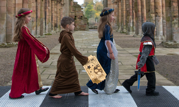 Image of a group of young kids walking across 'Abbey Road' dressed as medieval maidens and knights in the nave at Jedburgh Abbey