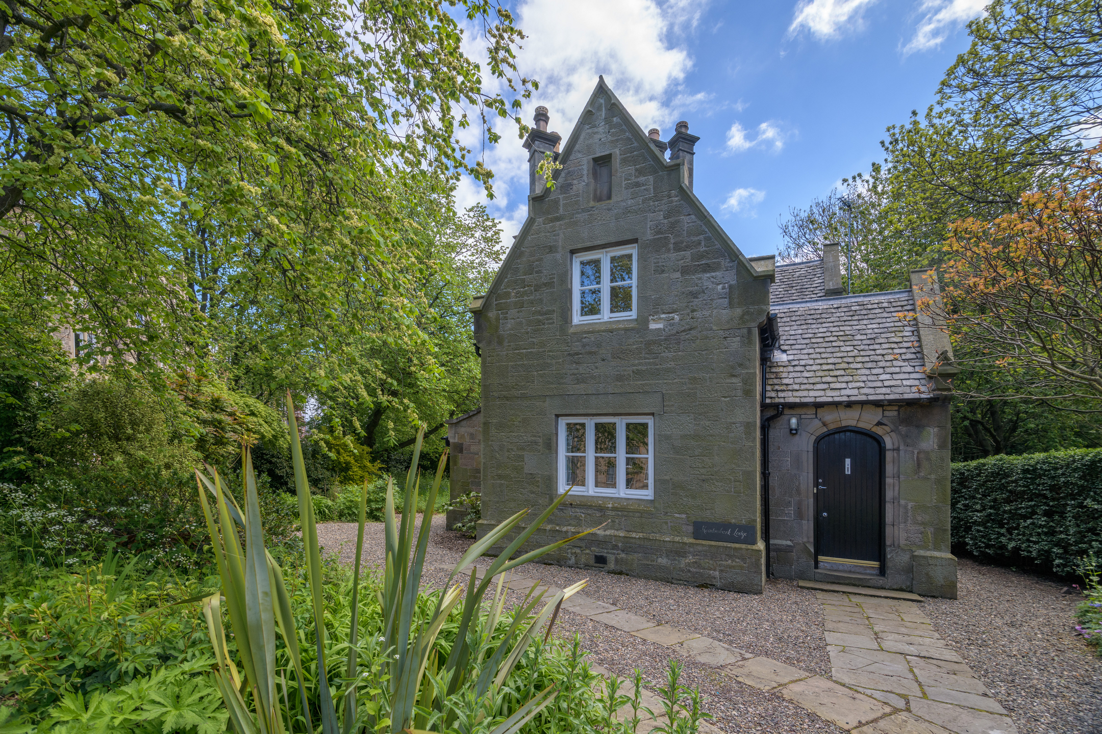 A large, stone lodge with a timber door and sash and case windows, in a lovely, green setting surrounded by trees and plants 