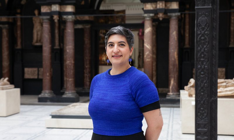 A person stands smiling and standing with their hands behind their back looking at the camera. They are wearing a blue jumper and standing in the foyer of a museum, with columns in the background.