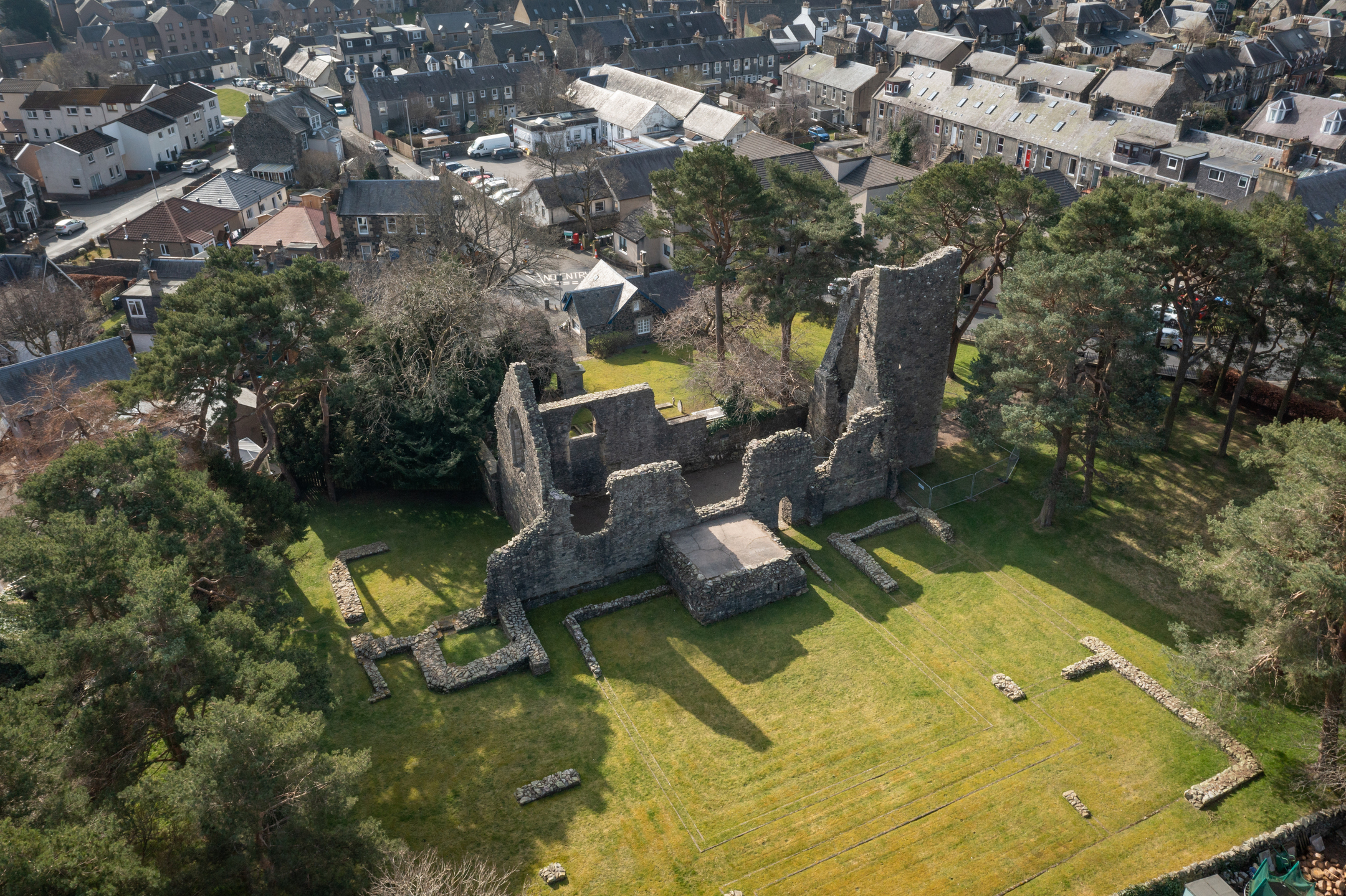 Aerial shot of Cross Kirk ruins in Peebles, showing the rectangular grassy site with stone foundations and surviving walls. The surrounding neighborhood of houses and gardens fills the frame.