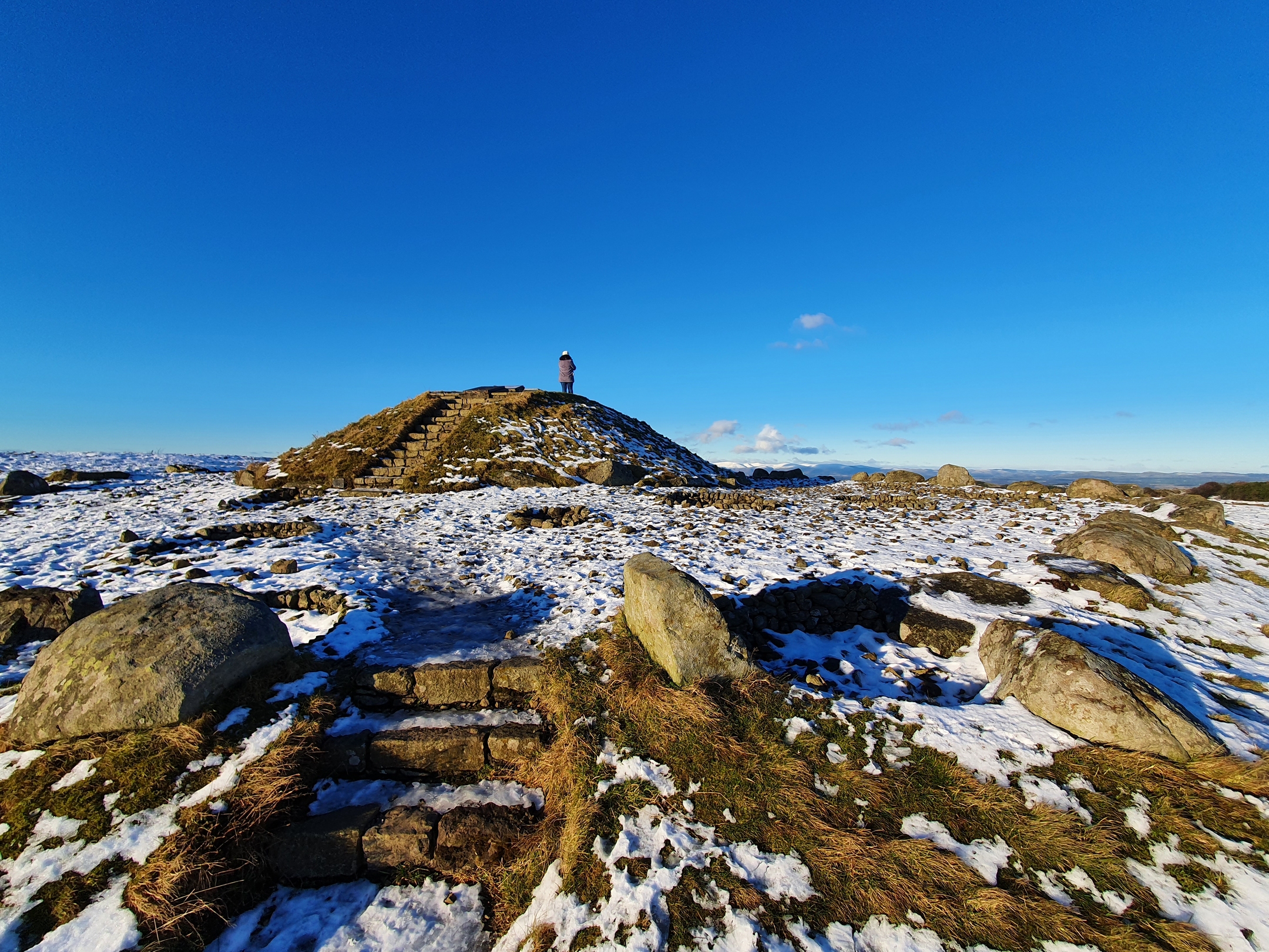 Small hill with person standing on top in grassy area with stones and light snow cover