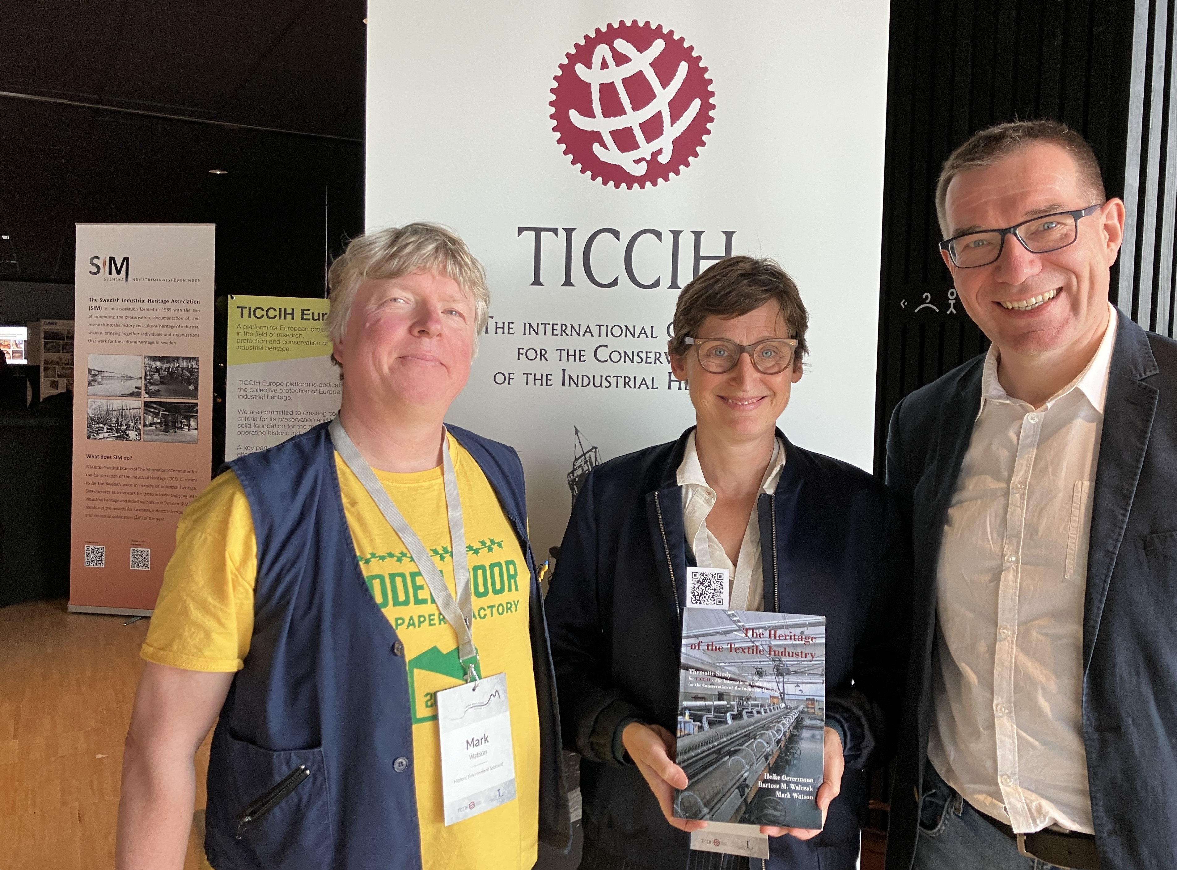 Three people standing indoors in front of a TICCIH banner; the person in the centre holds a book titled The Heritage of the Textile Industry. Exhibition banners and display stands are visible in the background.