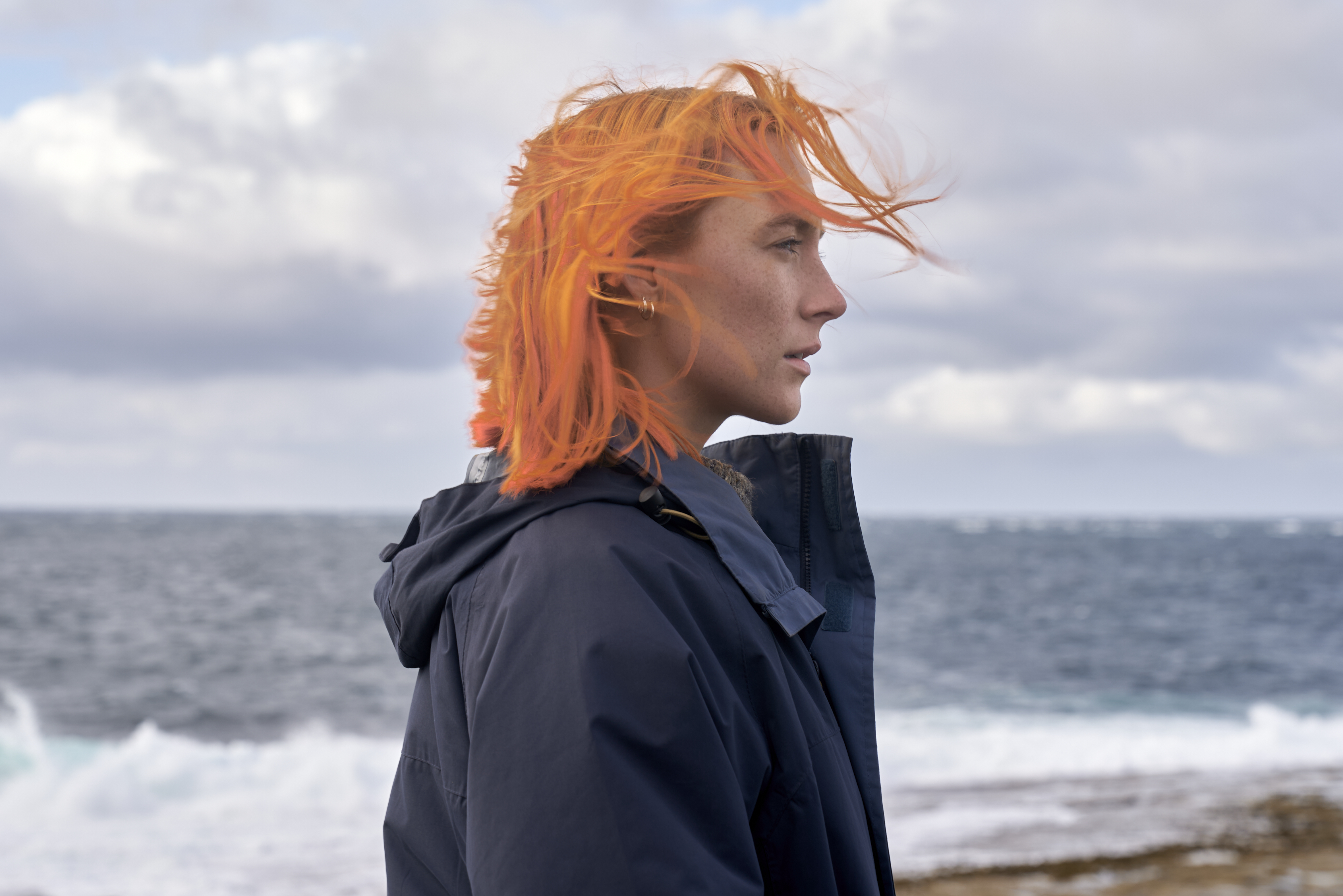 A woman with bright orange hair blowing in the wind stands by the sea, wearing a dark waterproof jacket. Waves crash behind her under a cloudy sky.