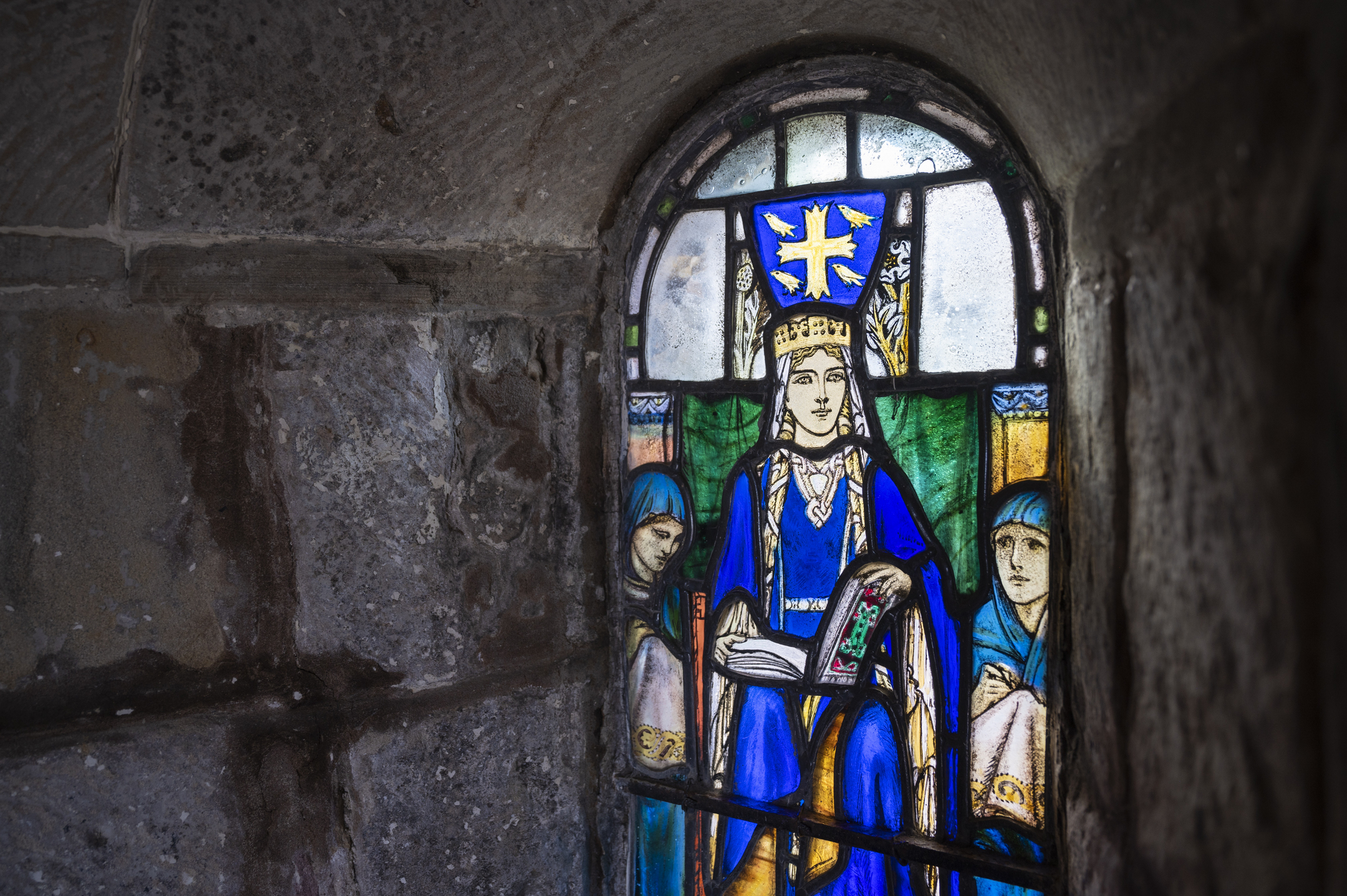 Stained-glass window showing a crowned seated figure holding an open book, flanked by smaller figures, set into a thick stone wall.