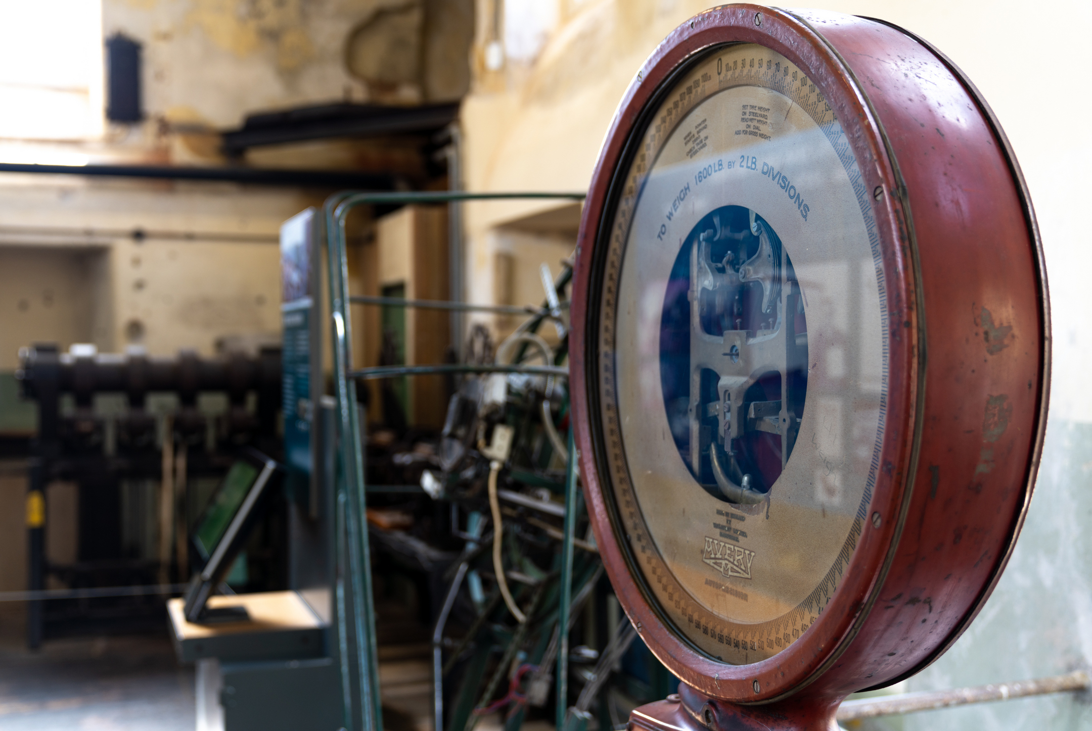 A close-up view of a large industrial weighing machine with a circular metal dial in a red casing. Behind it, blurred mill machinery sits in a worn, historic workshop interior.