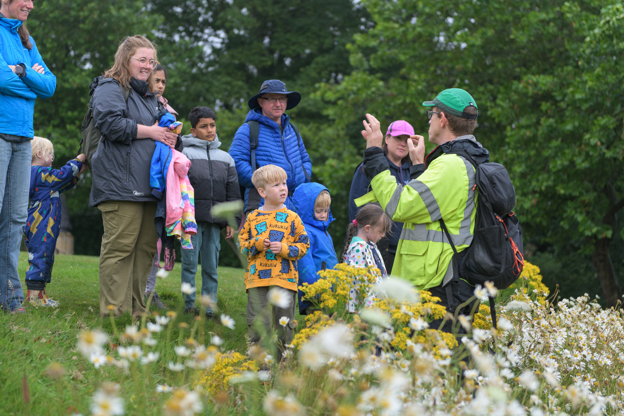 A group of adults and children gather on a grassy slope as an outdoor guide in a high‑visibility jacket gestures while talking, with wildflowers in the foreground and trees behind.