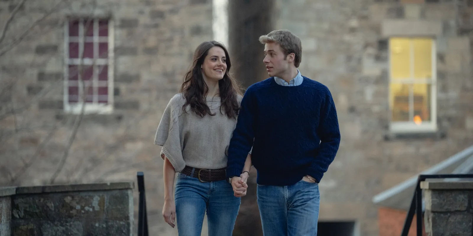 A young couple walks hand‑in‑hand outdoors near stone buildings. Both wear casual jumpers and jeans. They look at each other warmly as they walk along a path.