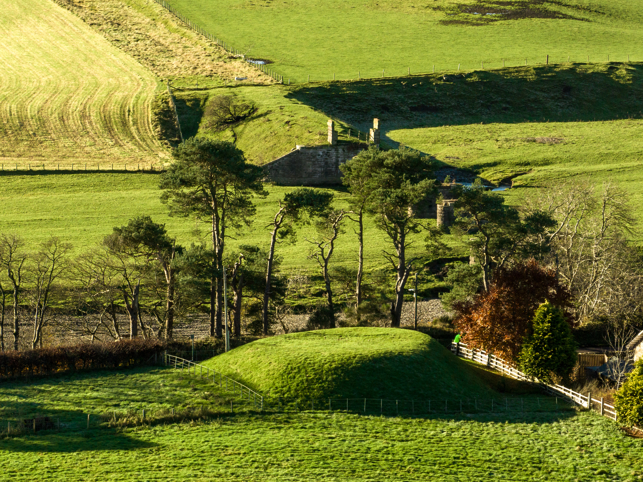 A grassy mound surrounded by trees. In the background are farmer's fields and the ruins of a former railway bridge. 
