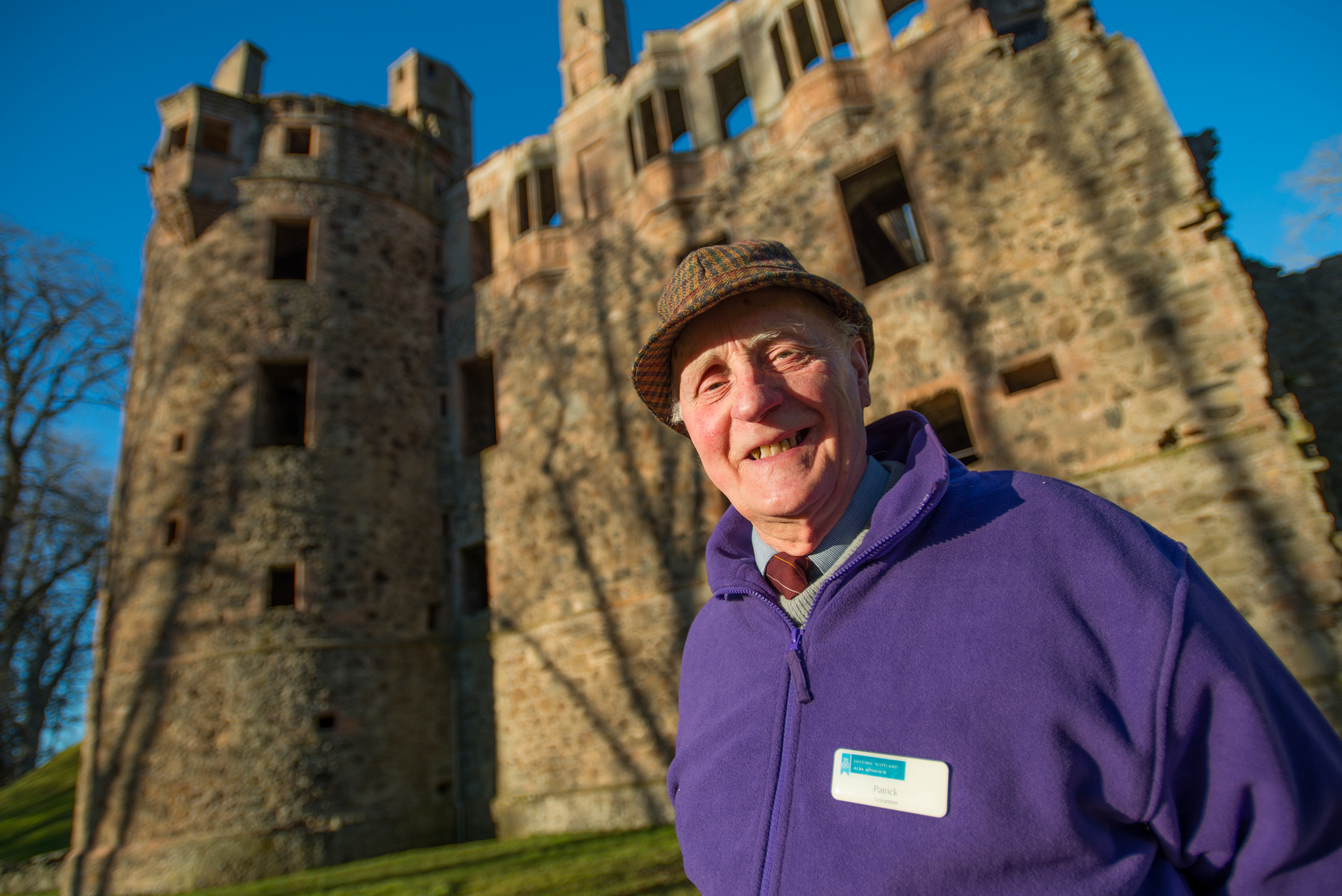 Close-up of a person wearing a purple fleece and flat cap standing in front of a historic stone castle with towers, under a clear blue sky.
