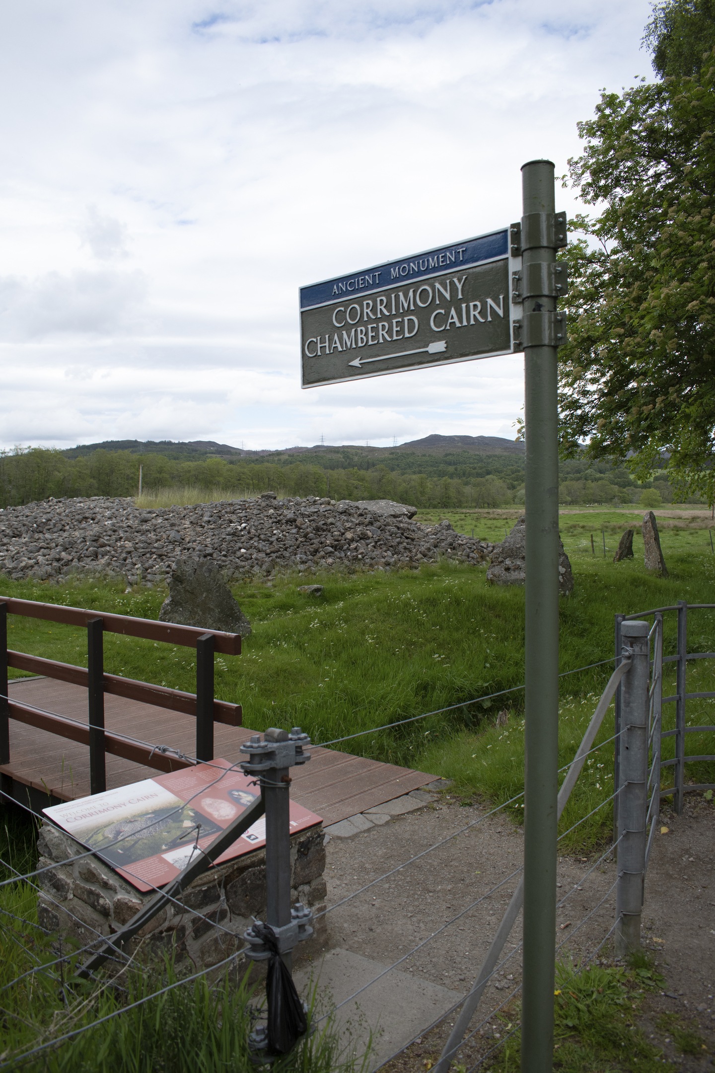 Signpost reading ‘Ancient Monument: Corrimony Chambered Cairn’ beside a small wooden bridge, with the stone cairn visible in the grassy field beyond.