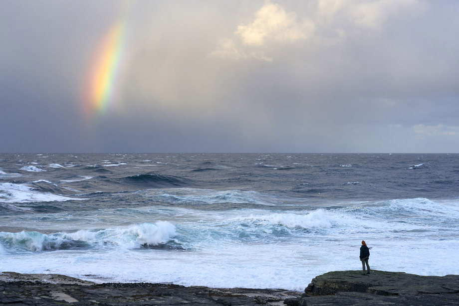 a woman standing on rocks looking out to a very choppy sea with a cloudy sky and bright firebow