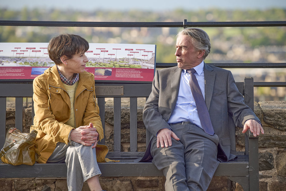 Left to right, actors Sally Hawkins & Steve Coogan sitting on a bench, with Edinburgh city centre in the background