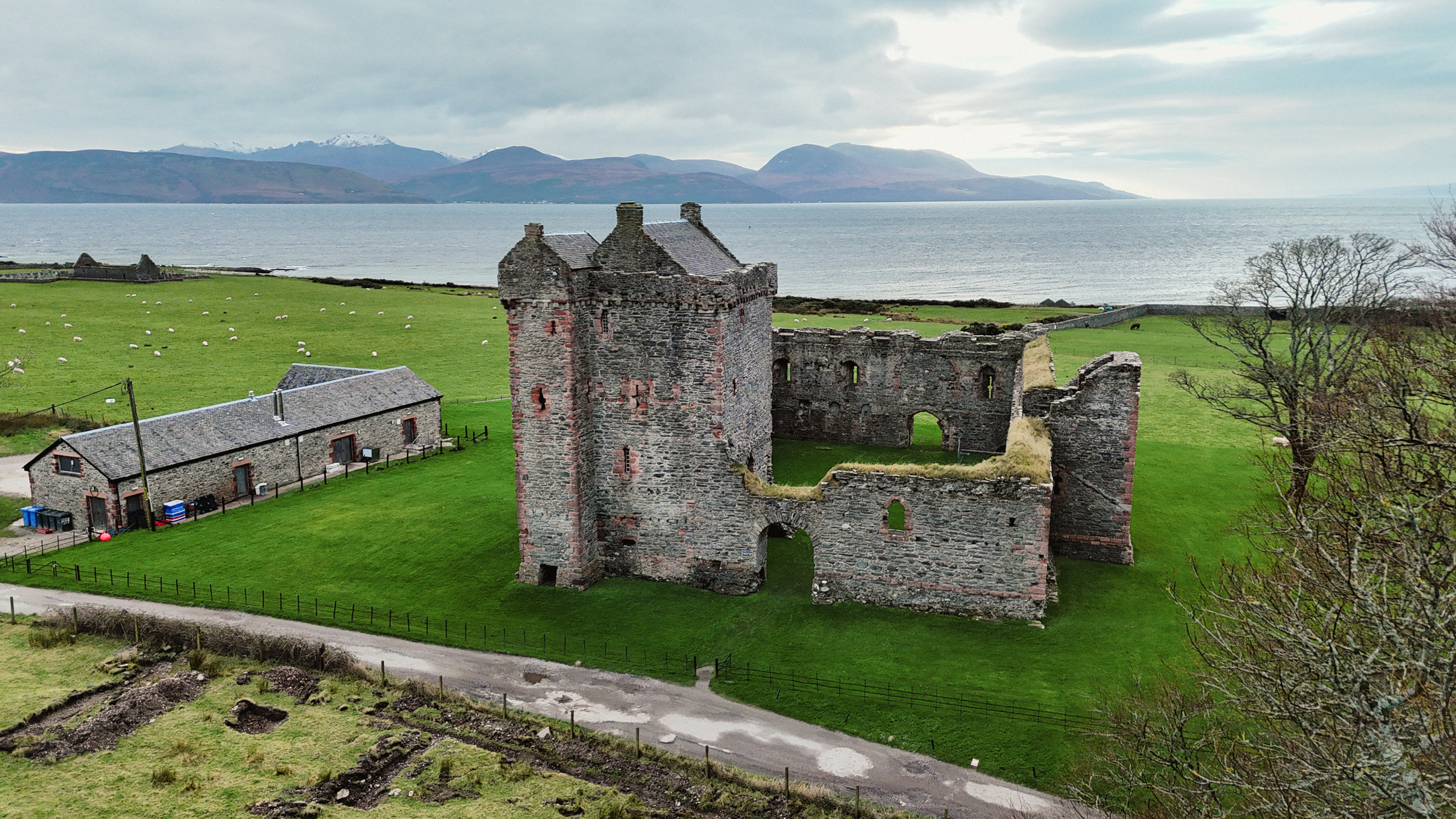 Drone view of a medieval stone castle ruin beside a small farm building, surrounded by green fields with the sea and distant hills in the background.