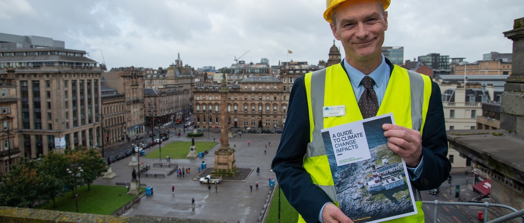 A person wearing a hardhat, standing on a balcony overlooking Glasgow's George Square, holding up a climate impacts guide