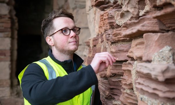 A person wearing glasses and a high vis vest inspects eroding castle rock
