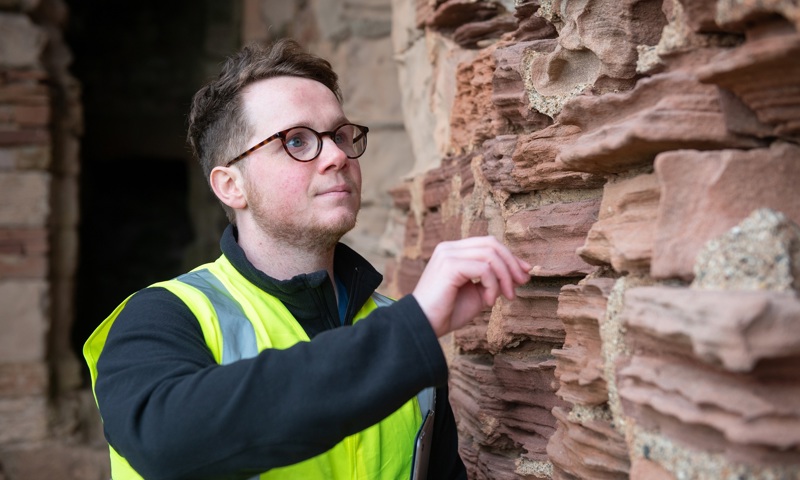 A person wearing glasses and a high vis vest inspects eroding castle rock