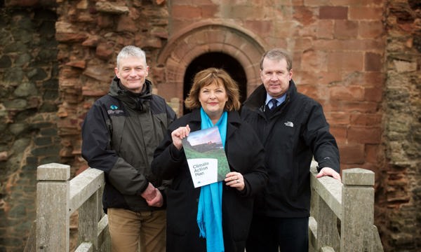 Three people stand at the entrance of a castle, one of them holding up a paper document