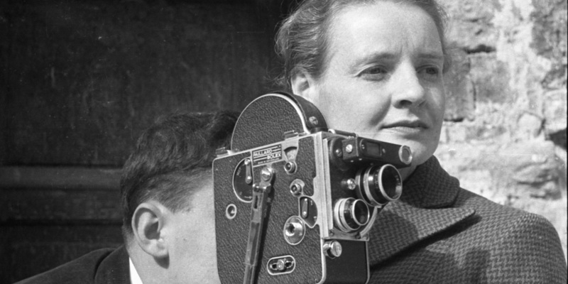 Black and white photo of a woman standing beside on old film camera. A man operates the camera.