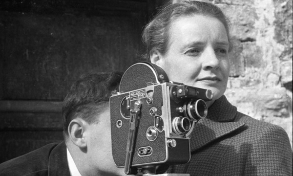 Black and white photo of a woman standing beside on old film camera. A man operates the camera.