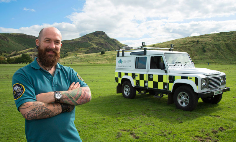 A man in a blue shirt with a beard stands in front of a large vehicle with a hill behind him.