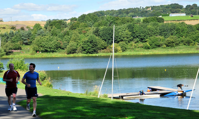 Two men running beside a loch on a sunny day