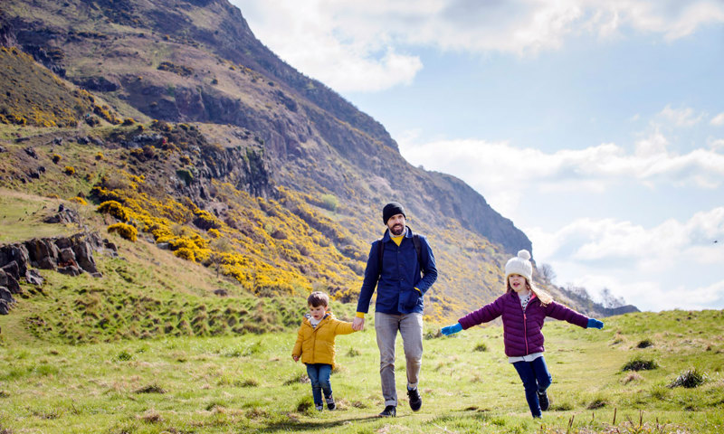A man in a hat walks with two children through a park with a hill covered in gorse in the distance